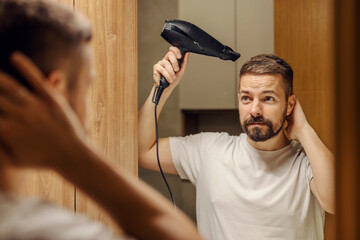A bearded man is looking himself in mirror reflection in a bathroom and drying his hair with...