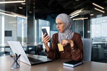 Upset and disappointed woman in hijab inside office trying to make purchase in online store, business woman holding smartphone and bank credit card, received money transfer error. © Liubomir