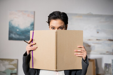 Short haired artist holding sketchbook near face in workshop