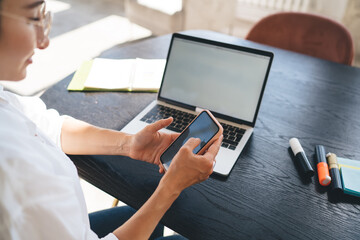 Crop woman using smartphone while working on laptop