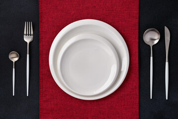 Festive place setting with red napkin. Empty plates and silver cutlery on dark black background....