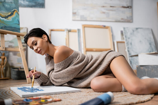 Sexy short haired artist painting on canvas on floor in blurred workshop