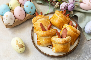 Buns in the form of an Easter rabbit from sausage and cheese in a yeast dough with colored eggs on a gray stone background. Traditional Easter symbols and food.
