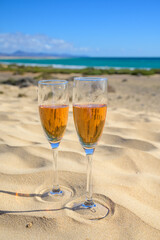 Two glasses of rose champagne or cava sparkling wine served on white sandy tropical beach with dunes and blue ocean, romantic vacation, winter sun on Fuerteventura, Canary, Spain