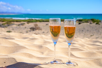 Two glasses of rose champagne or cava sparkling wine served on white sandy tropical beach with dunes and blue ocean, romantic vacation, winter sun on Fuerteventura, Canary, Spain