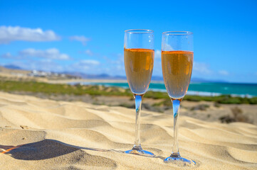 Two glasses of rose champagne or cava sparkling wine served on white sandy tropical beach with dunes and blue ocean, romantic vacation, winter sun on Fuerteventura, Canary, Spain