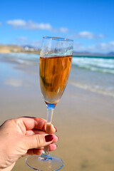 Hand with glass of rose champagne or cava sparkling wine with view on white sandy tropical beach and blue ocean water, romantic vacation, winter sun on Fuerteventura, Canary, Spain