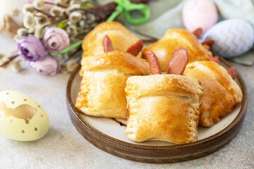 Traditional Easter symbols and food. Buns in the form of an Easter rabbit from sausage and cheese in a yeast dough with colored eggs on a gray stone background.