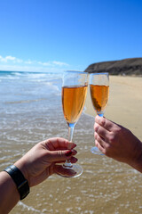Beach party, hands with glass of rose champagne or cava sparkling wine, view on white sandy tropical beach and blue ocean water, romantic vacation, winter sun on Fuerteventura, Canary, Spain