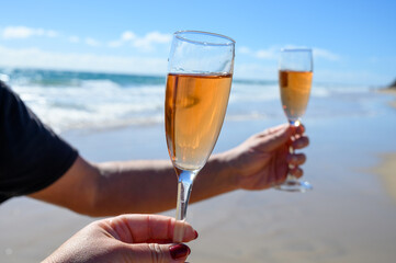 Beach party, hands with glass of rose champagne or cava sparkling wine, view on white sandy tropical beach and blue ocean water, romantic vacation, winter sun on Fuerteventura, Canary, Spain