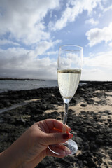 Hand with glass of white champagne or cava sparkling wine on white sandy tropical beach and blue ocean, romantic vacation, winter sun on Fuerteventura, Canary, Spain