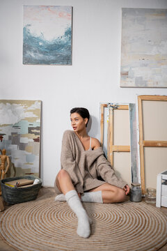 Short haired artist in sweater sitting near paintings on floor in workshop