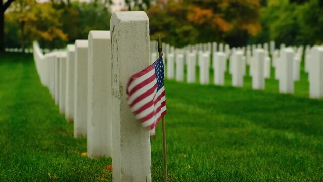 Second World War Cemetery. Military Cemetery Decorated for Memorial Day. American flag with stripes and stars flutters in strong wind. Cemetery graveyard white tombstones at sunset sky 
