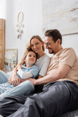 cheerful woman looking at camera near happy husband and daughter in bedroom.