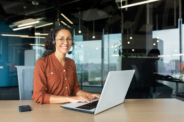 Fototapeta na wymiar Portrait of Latin American business woman, office worker looking at camera and smiling, using headset and laptop for remote online communication, customer support tech call center worker.