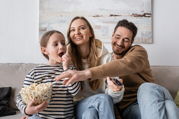 kid looking at happy father taking popcorn near cheerful mother with remote controller.