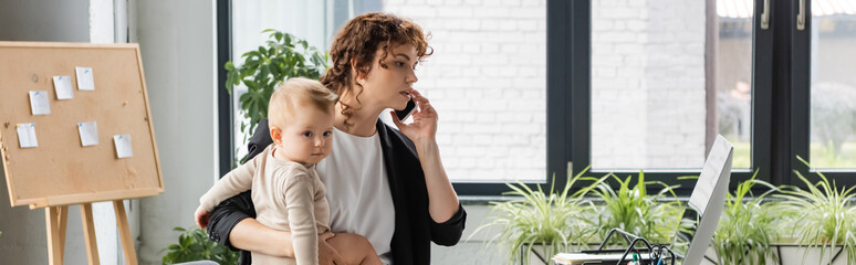 Fototapeta premium businesswoman talking on mobile phone and holding little daughter near computer monitor in office, banner.