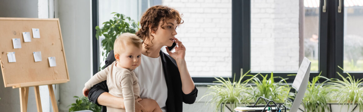 businesswoman talking on mobile phone and holding little daughter near computer monitor in office, banner.