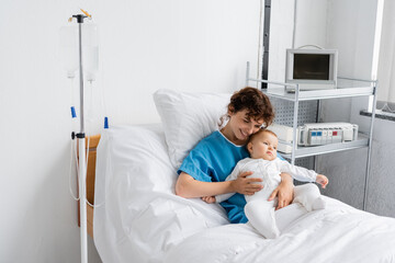 Obraz premium joyful woman in patient gown hugging little baby in romper while sitting on hospital bed.