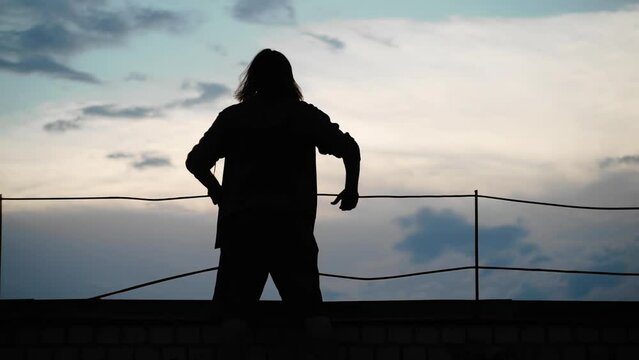 A guy standing on the roof of a house at sunset and the wind blows over him. his silhouette is clearly against the background of the evening sky