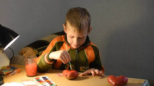 The boy paints with watercolors an object lying in front of him on the table.