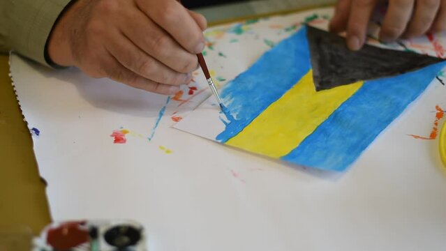 The artist is sitting at the table. He draws the flag of the Bahamas with a brush and paints.