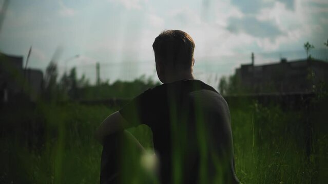 The guy is sitting in the grass at the edge of the path outside the city. The camera shoots through the grass from behind. The atmosphere of loneliness