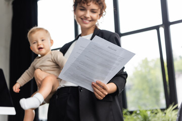 cheerful businesswoman standing with toddler child and looking at documents in office.