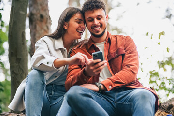 A couple is watching something funny on their smart phone during their rest after nature exploration and hiking, they look content and happy.
