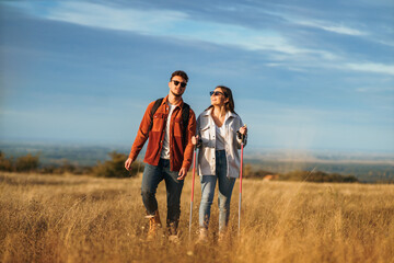 A couple is chatting and hiking in big golden yellow meadow on a sunny autumn day. They wear sunglasses to protect them from sun. The girl is holding trekking poles to help her while hiking.