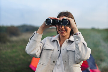 A girl is holding binoculars with both hands to look at a distance standing in a green meadow.