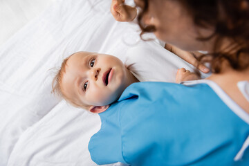 top view of little child looking away while lying on hospital bed near blurred mom.