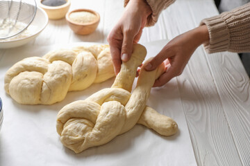 Woman cooking braided bread at white wooden table in kitchen, closeup. Traditional Shabbat challah - obrazy, fototapety, plakaty