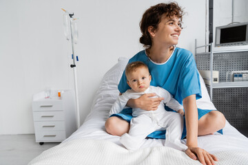 cheerful woman in patient gown looking away while sitting with toddler baby on hospital bed.