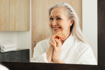 Aged woman admires her reflection in a bathroom mirror. Smiling senior female in a bathrobe.