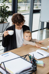 businesswoman sitting with little daughter and coffee cup near documents and blank notebook in office.