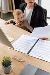 Fototapeta na wymiar blurred businesswoman holding document while sitting at work desk with toddler daughter.