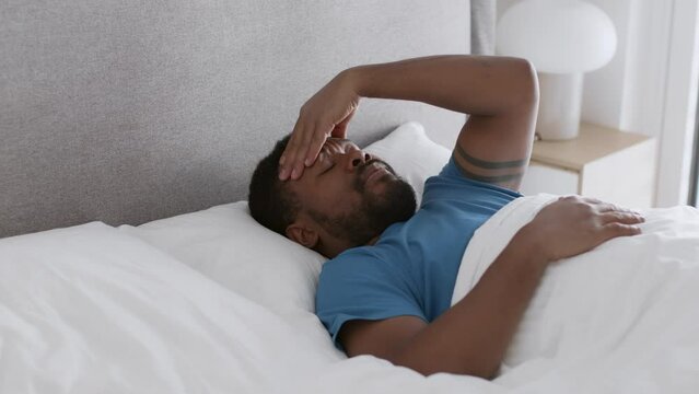 Bad sleep. Young african american guy waking up in bed and massaging forehead, suffering from headache and keep resting