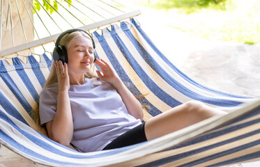 A young blonde girl lies in a hammock and listens to music in wireless headphones