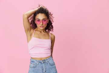 Happy woman hands up waxing her armpits, shaving, with curly hair in a pink tank top and jeans on a pink background wearing sunglasses with a nice tan, copy space