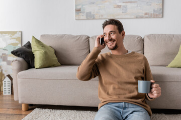 cheerful man in beige jumper talking on smartphone while sitting with cup of coffee at home.