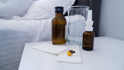 Medicine, pills, bottle of syrup, nasal spray and a glass of water on the white bedside table. Illness treatment