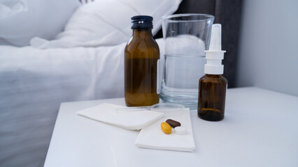 Medicine, pills, bottle of syrup, nasal spray and a glass of water on the white bedside table....