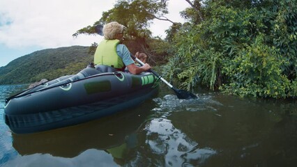 Granny with kid kayaking. Elderly women paddles inflatable kayak with toddler girl on the calm freshwater lake. Family explore the coast with lush trees and mangroves on kayak