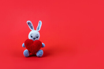 Knitted gray bunny with a crocheted red heart on a red background. Happy Valentine's Day, Mother's Day and birthday greeting card.