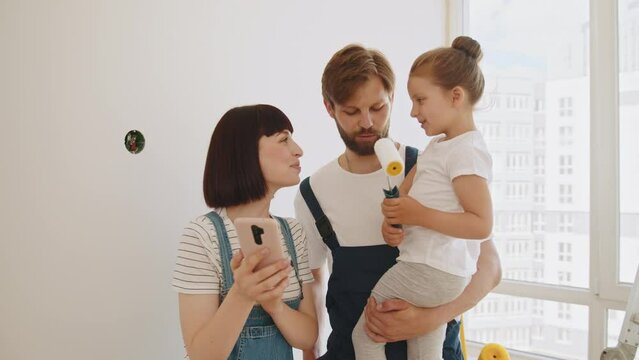 Home repair. A cheerful happy family is renovating an apartment. Joyful young caring parents with little cute preschool girl, tapping and browsing online on smartphone, choosing new room decor
