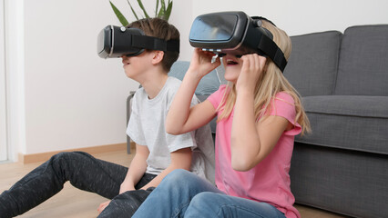 Two happy Caucasian siblings sitting in the living room, putting on VR headsets and enjoying a 3d experience. Indoors gaming, fun, activity concept.