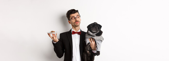 Handsome young dog owner in fancy suit holding cute black pug and plate with animal food, standing...