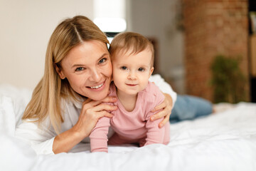 Happy motherhood. Mommy playing with adorable toddler baby crawling on bed, smiling at camera, bedroom interior