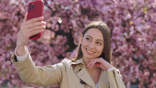 Cheerful young woman in beige trench coat using modern smartphone for taking selfie near blooming sakura. Modern gadgets for saving happy moments